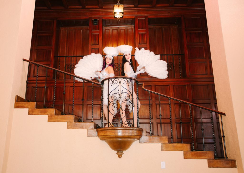 Showgirls in white and silver costumes posing with feather fans. On a staircase with a dark wood background.